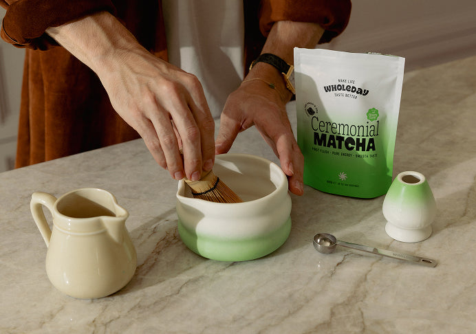 A Wholeday matcha pouch with a latte, a bamboo whisk in matcha powder, and an iced matcha in hand
